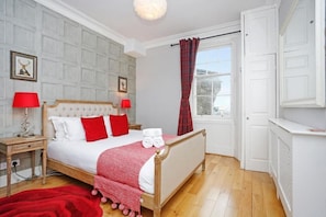 Une chambre à coucher bien rangée, avec un grand lit, deux tables de chevet, un plaid rouge et une fenêtre avec des rideaux.