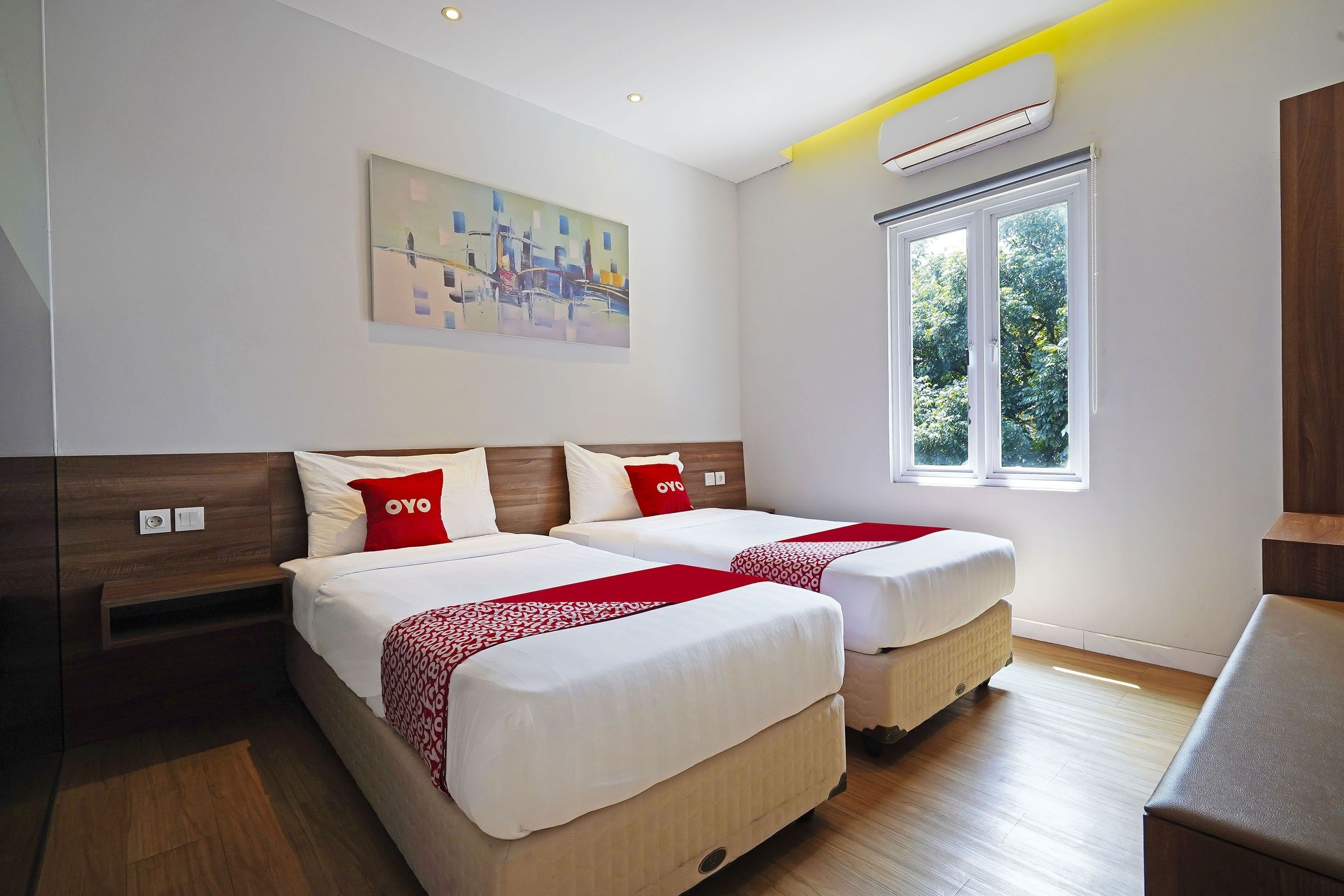 A hotel room with two beds, a painting on the wall, a window with a view of trees, and an air conditioning unit.