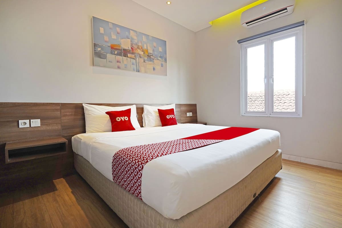 Une chambre d’hôtel moderne avec un grand lit, un couvre-lit aux motifs rouges et blancs, et un tableau représentant un paysage urbain accroché au mur.