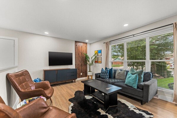 A modern living room with a leather armchair, a sofa with decorative pillows, a flat-screen TV mounted on the wall, and a large window with blinds. - Spacious 5-Bed Home with Hot Tub (Denver)