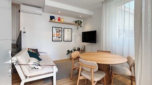 Een moderne woonkamer met een bank, een ronde houten eettafel en een flatscreen televisie aan de muur.