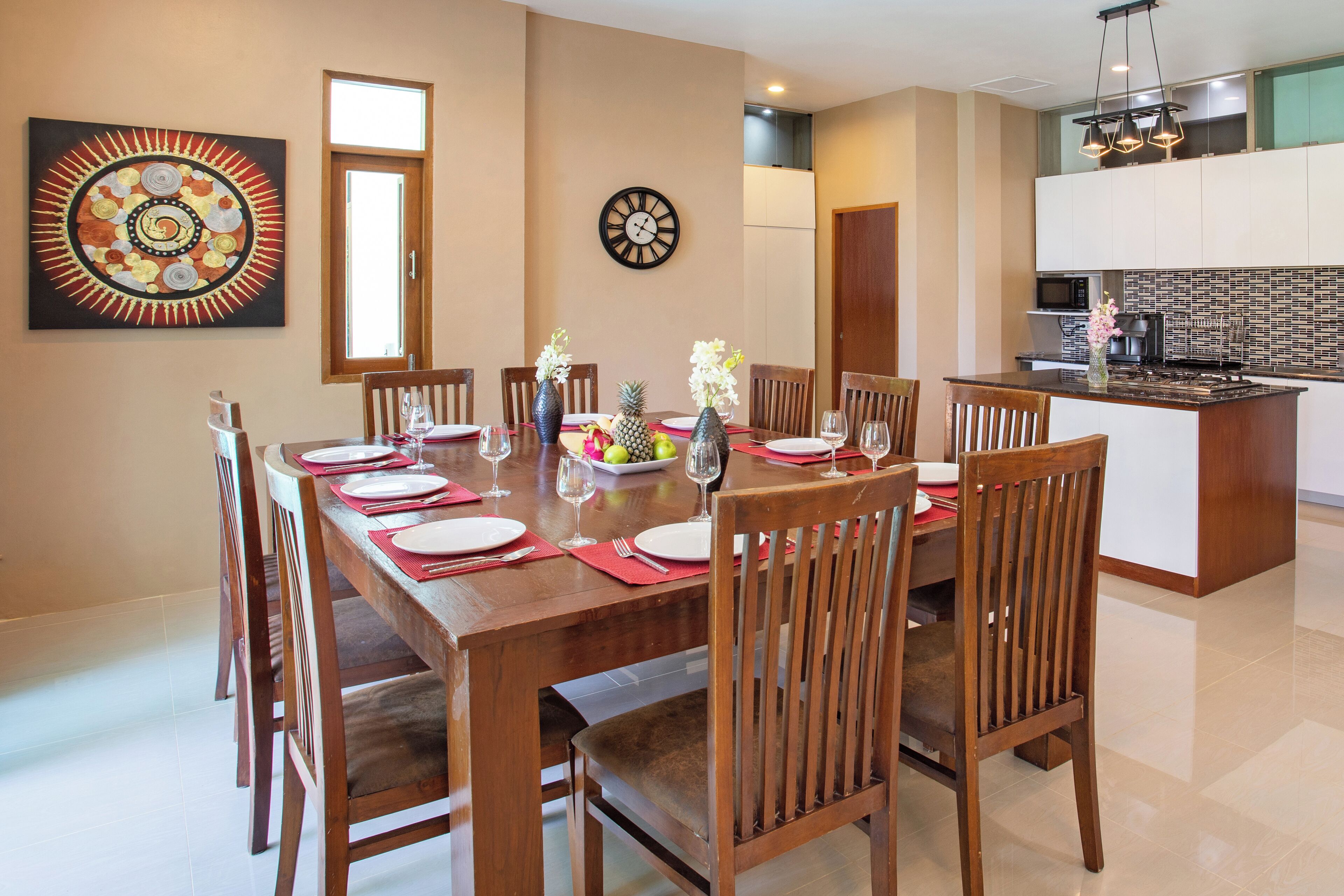 A dining area with a wooden table set for six, surrounded by chairs. A clock and a painting are on the wall.