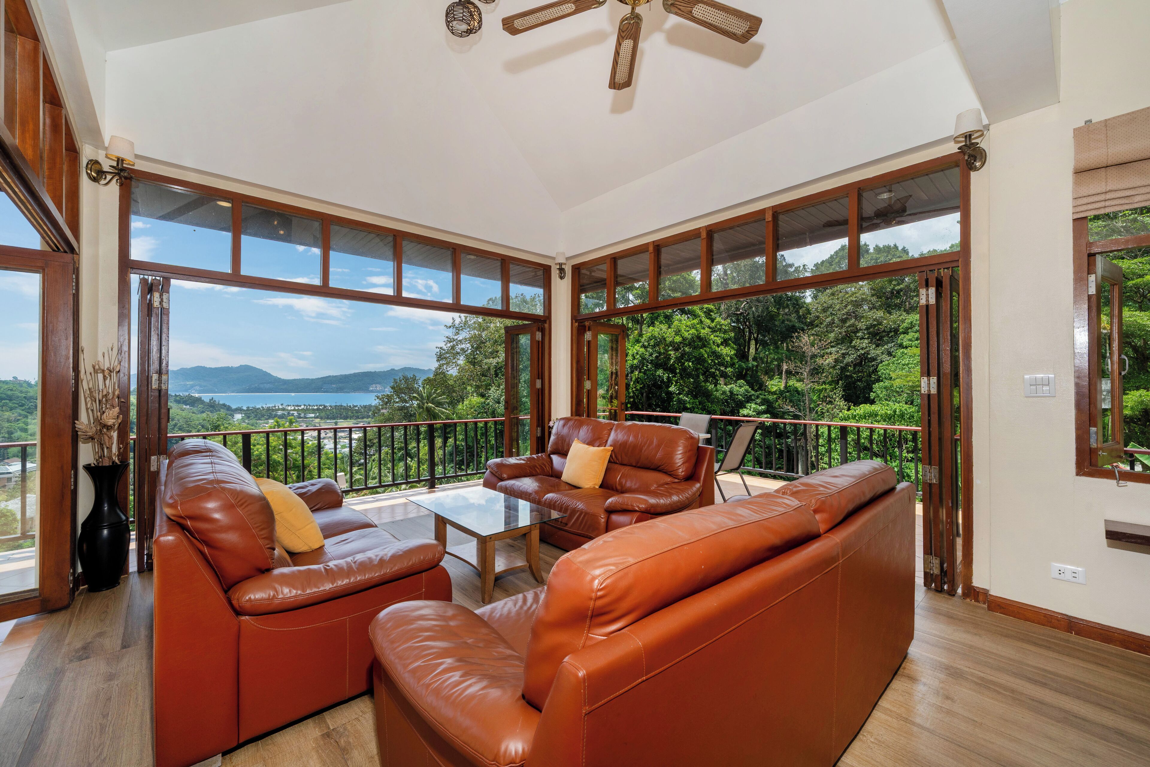 A spacious balcony with leather sofas and a table, large windows offering a view of greenery and mountains.