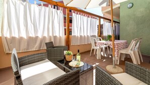 En udendørs terrasse med fletmøbler, et bord med en flaske, og et overdækket område med stole og bord.