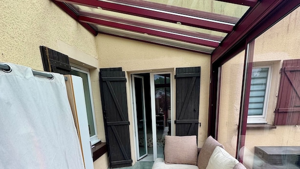 Une terrasse ensoleillée avec un canapé beige, une petite table ronde et une vue sur une maison à travers des volets ouverts.