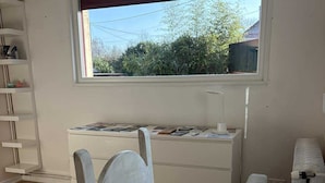 Une pièce avec une table blanche, une chaise blanche, une fenêtre donnant sur un paysage verdoyant et une étagère blanche sur laquelle sont posés divers objets.