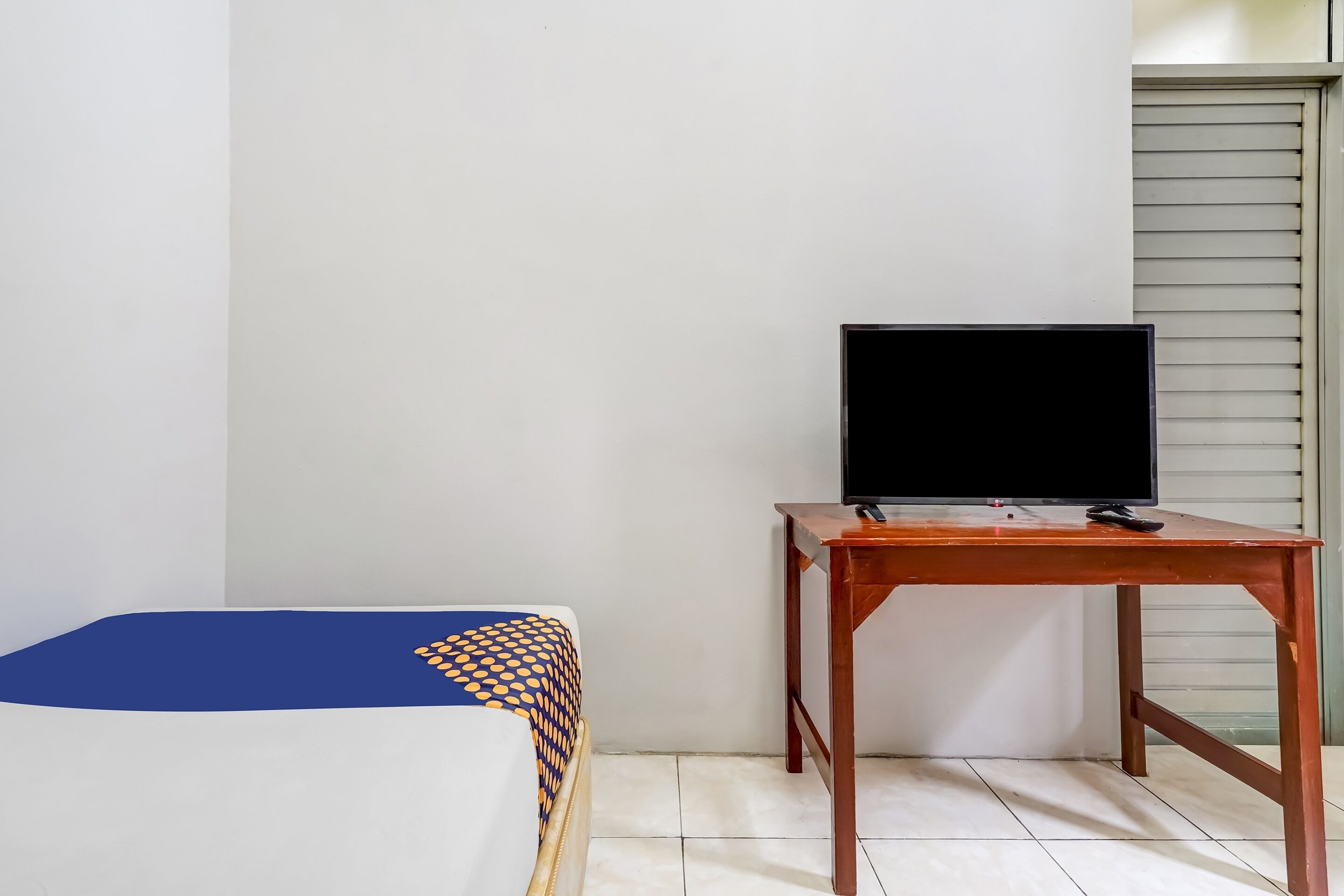 A room with a bed, a wooden table with a television, and a window with blinds.