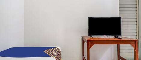 A room with a bed, a wooden table with a television, and a window with blinds.