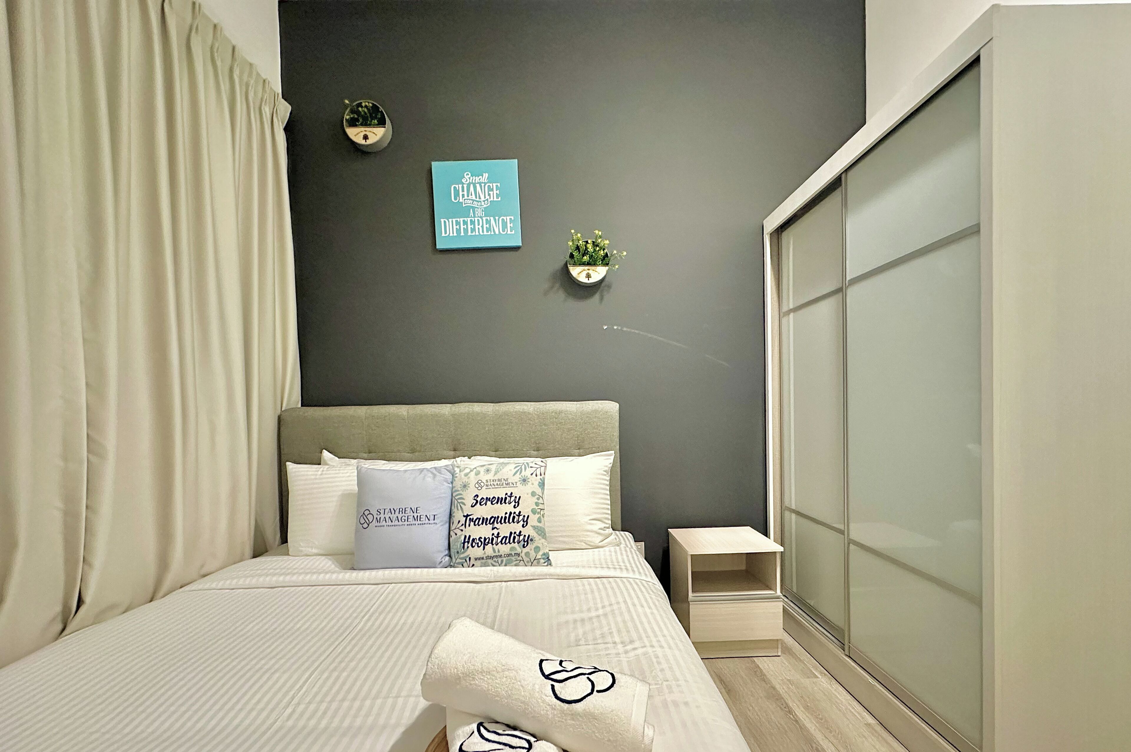 Una cama bien hecha con edredón blanco y almohadas, una mesita de noche, un reloj de pared, una cita inspiradora en un marco y una planta en maceta.