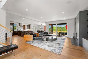 A spacious living area with a large sofa, a coffee table, and a fireplace. There is a kitchen in the background with modern appliances and a dining area.