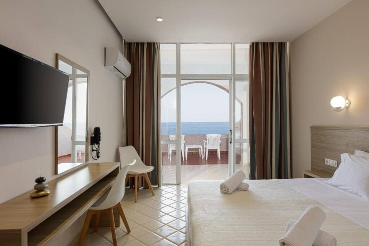 Habitación de hotel con cama, escritorio, televisor, teléfono y balcón con vista al mar.