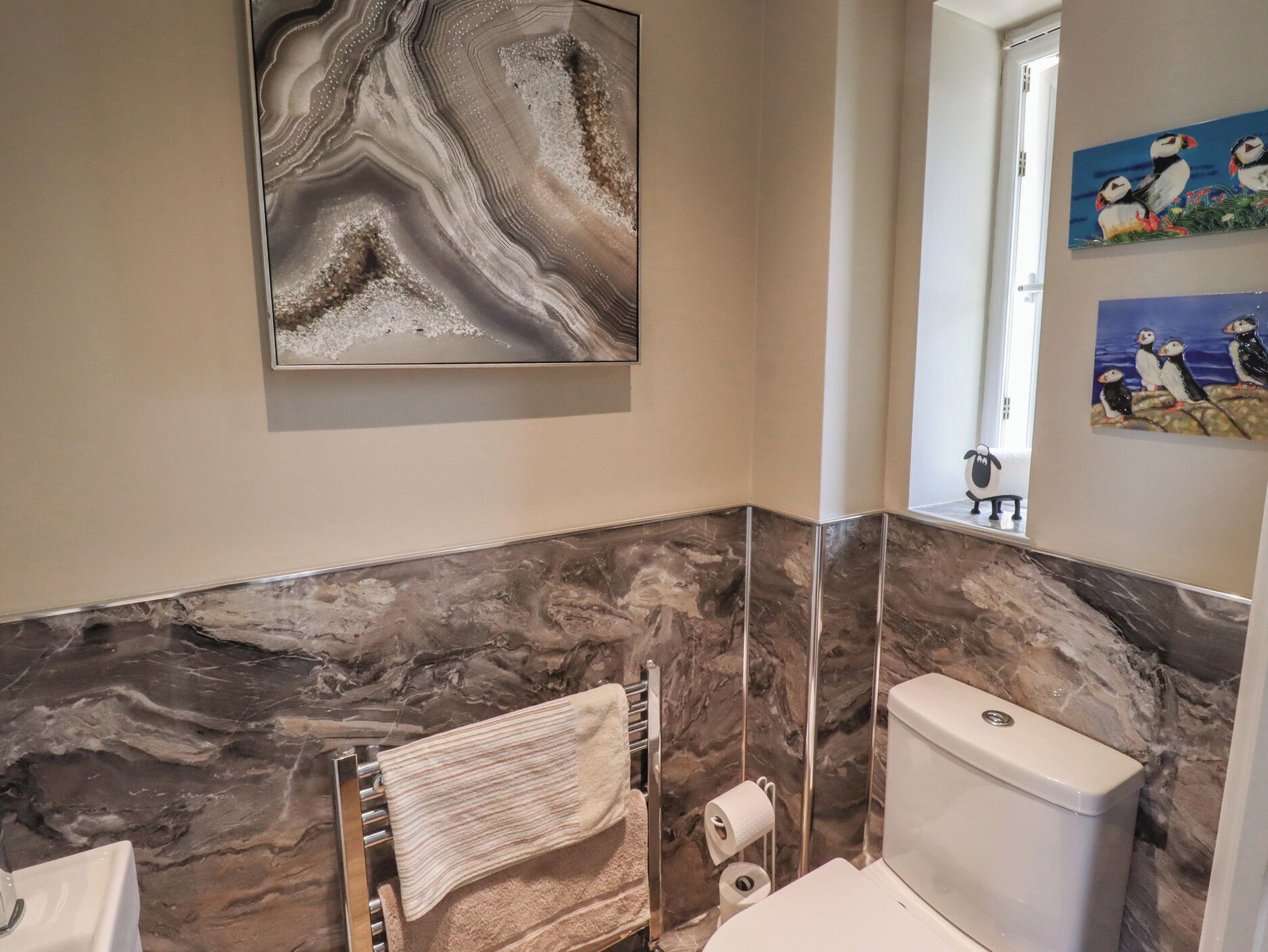 A bathroom with marble walls, a toilet, a towel rack, and a painting of puffins.