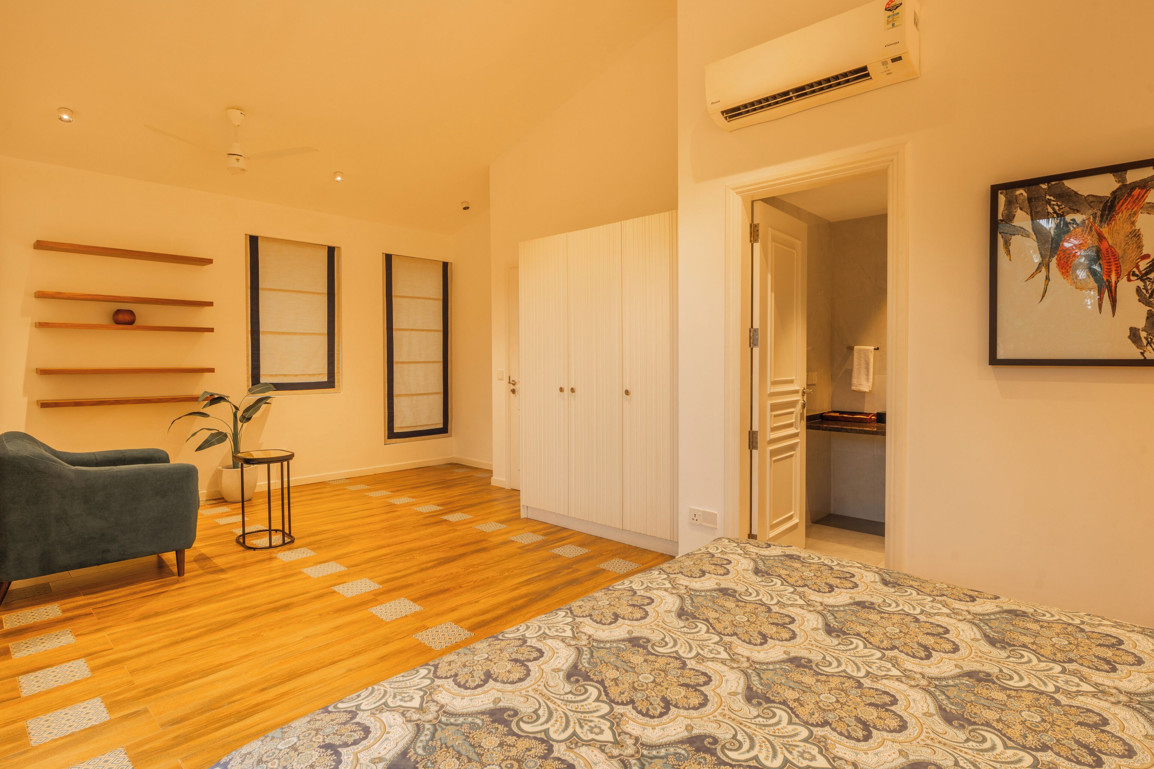 A modern bedroom with wooden flooring, a patterned bedspread, a grey armchair, a small side table, and a wardrobe.