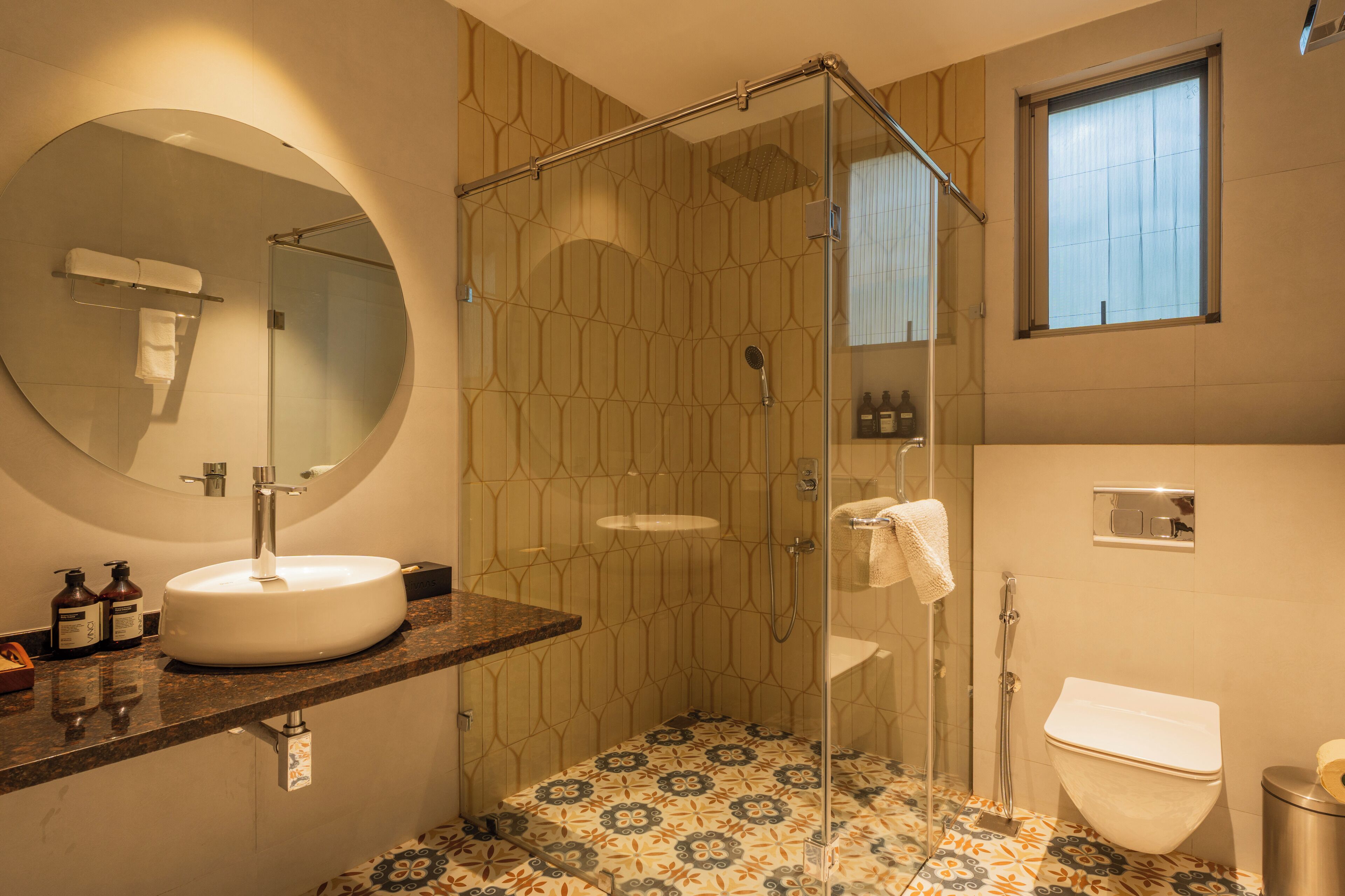 A bathroom with a glass-enclosed shower, a round mirror, a sink with a marble countertop, and patterned floor tiles.