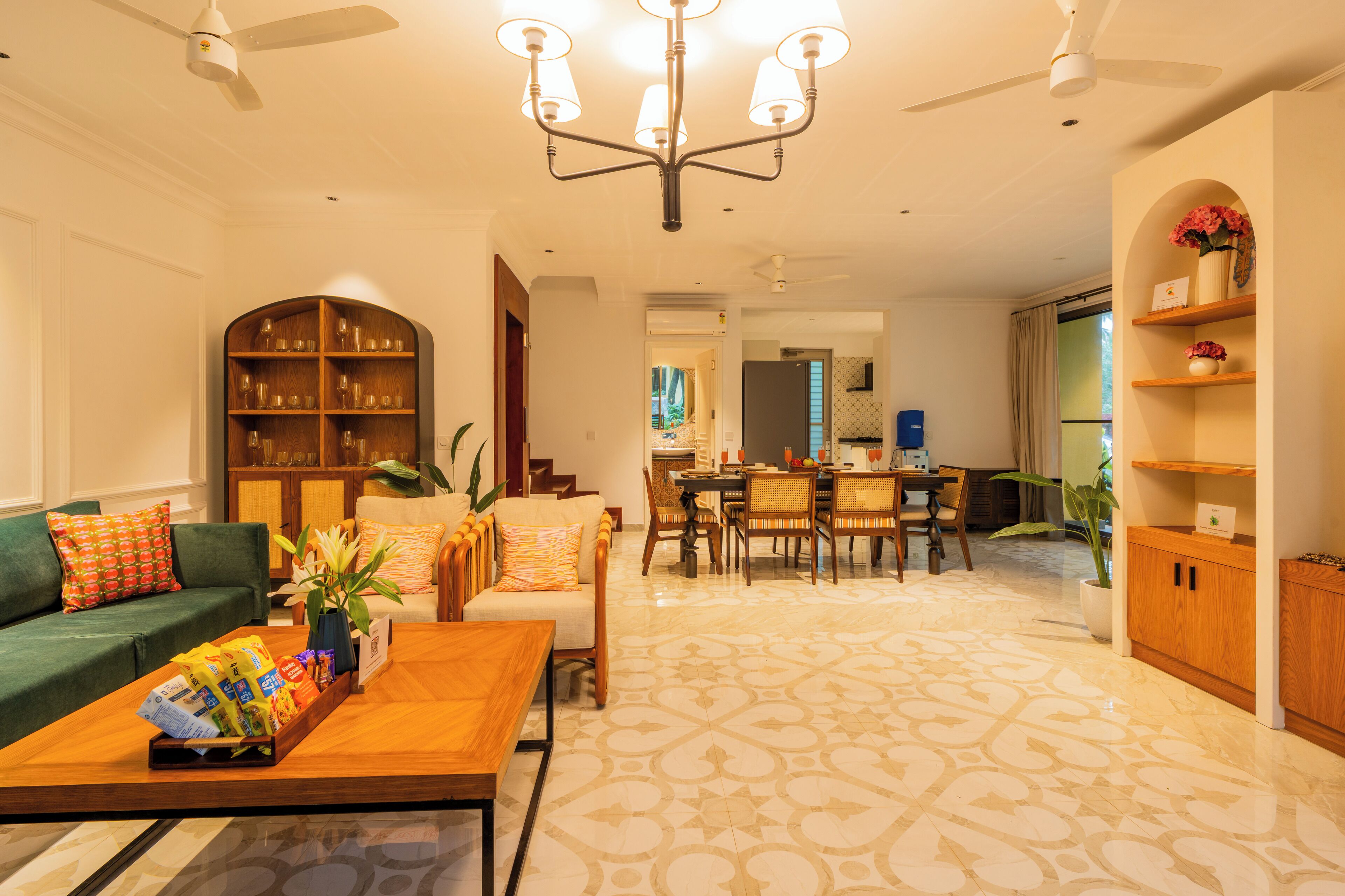 A spacious living room with a green sofa, a wooden coffee table with snacks, a chandelier, and a dining area with a table and chairs.