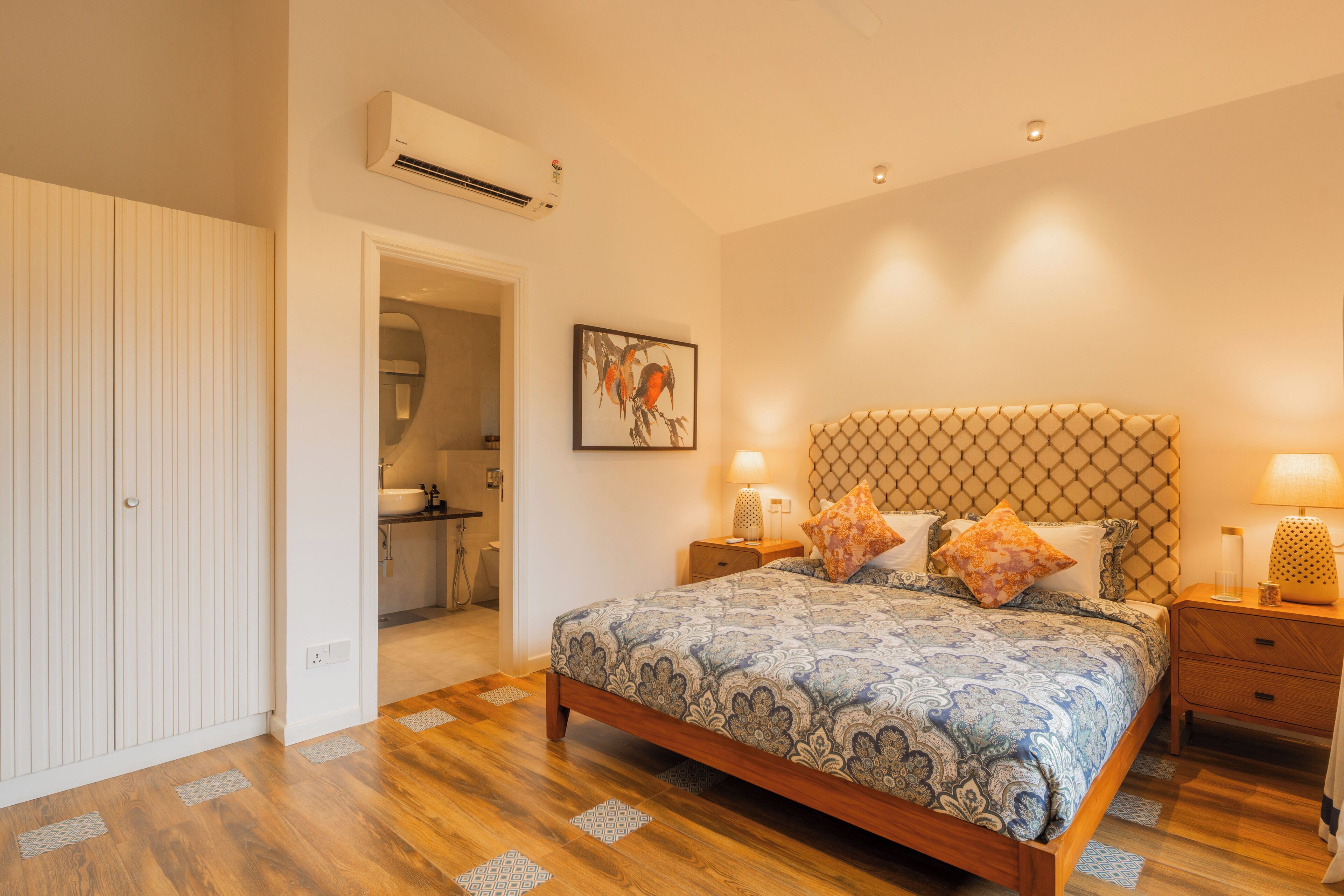Une chambre à coucher avec un lit en bois, une tête de lit ornée d’un motif géométrique, deux tables de chevet avec des lampes et un climatiseur fixé au mur.