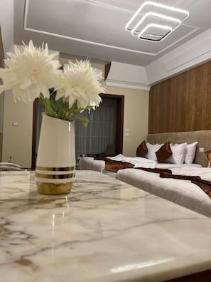Une chambre d’hôtel avec une table de chevet en marbre, un vase contenant des fleurs blanches, un lit avec des oreillers blancs et bruns, et une tête de lit en bois.