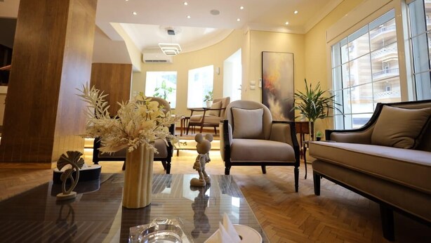 Un salon moderne avec un canapé, des fauteuils et une table basse. Une grande fenêtre laisse entrer la lumière naturelle et offre une vue sur le bâtiment situé à l’extérieur.