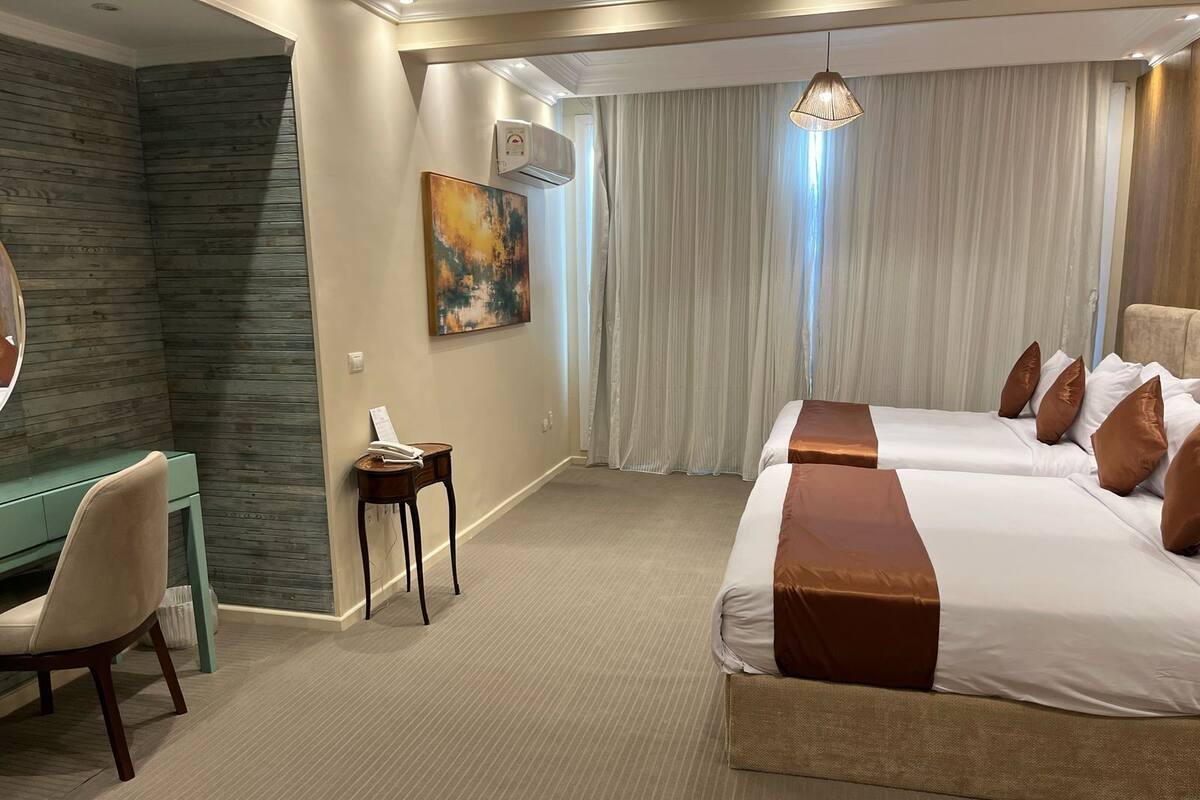 A hotel room with a desk, two beds, a painting, and a large window with curtains.