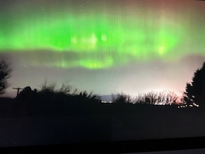 A view of the Northern Lights over a landscape with silhouettes of trees and a distant structure.