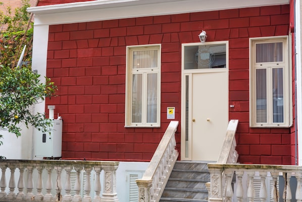 Un edificio de ladrillo rojo con una puerta blanca, dos ventanas y un pequeño balcón con barandilla.
