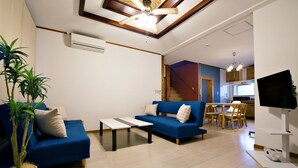 A modern living room with a blue sofa, a coffee table, and a potted plant. There is a staircase leading to a kitchen area with wooden cabinets and a dining table. - Onnason Yamada House (Onna)