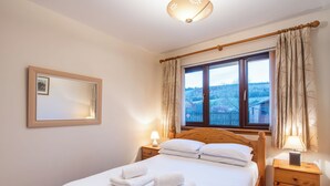 Una cama bien hecha con sábanas y toallas blancas, una lámpara de noche, una mesita de madera, un cuadro en la pared y una ventana con cortinas.
