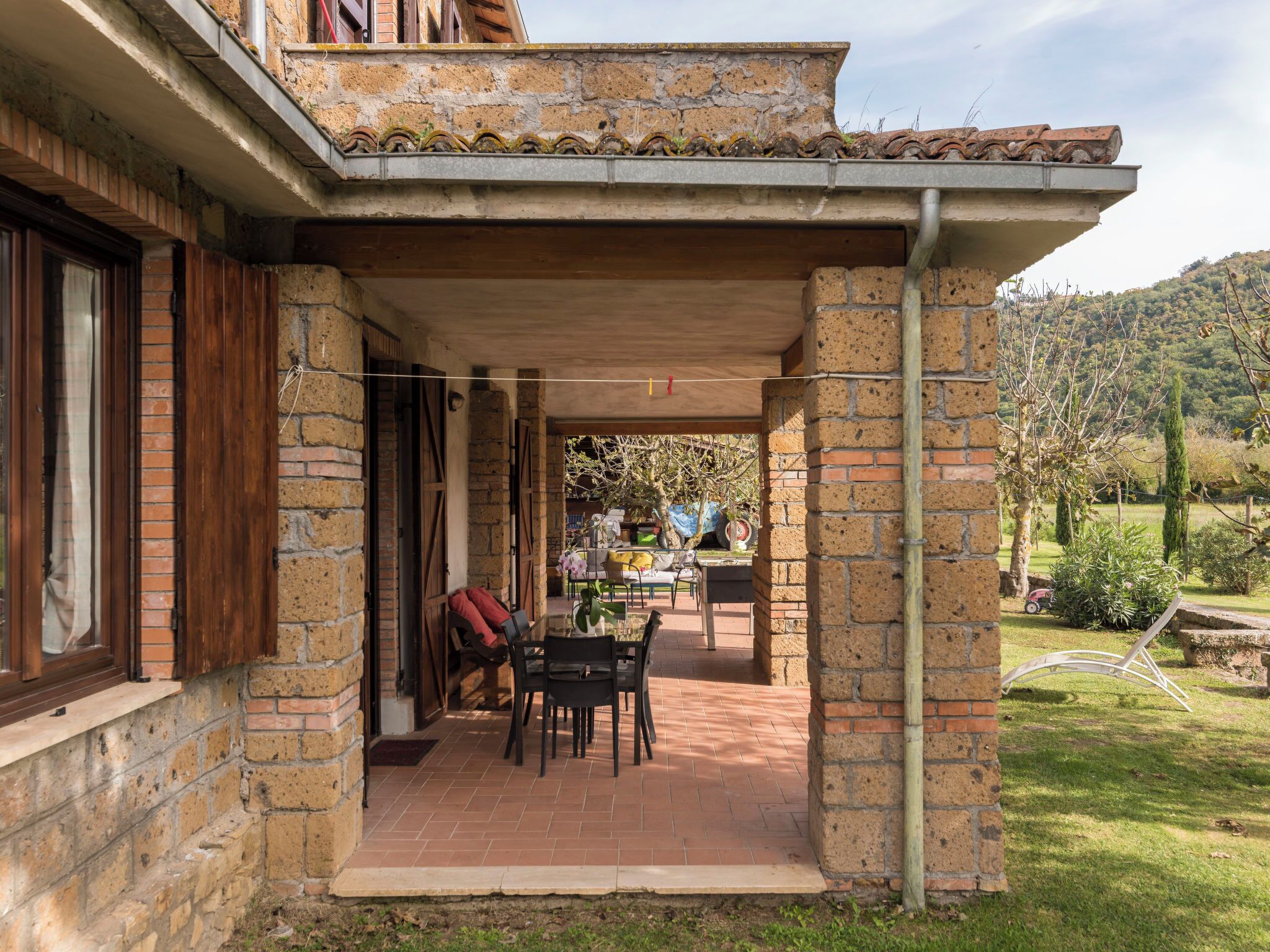 A covered patio area with brick walls, a wooden table and chairs, and a view of a garden and hills in the background.