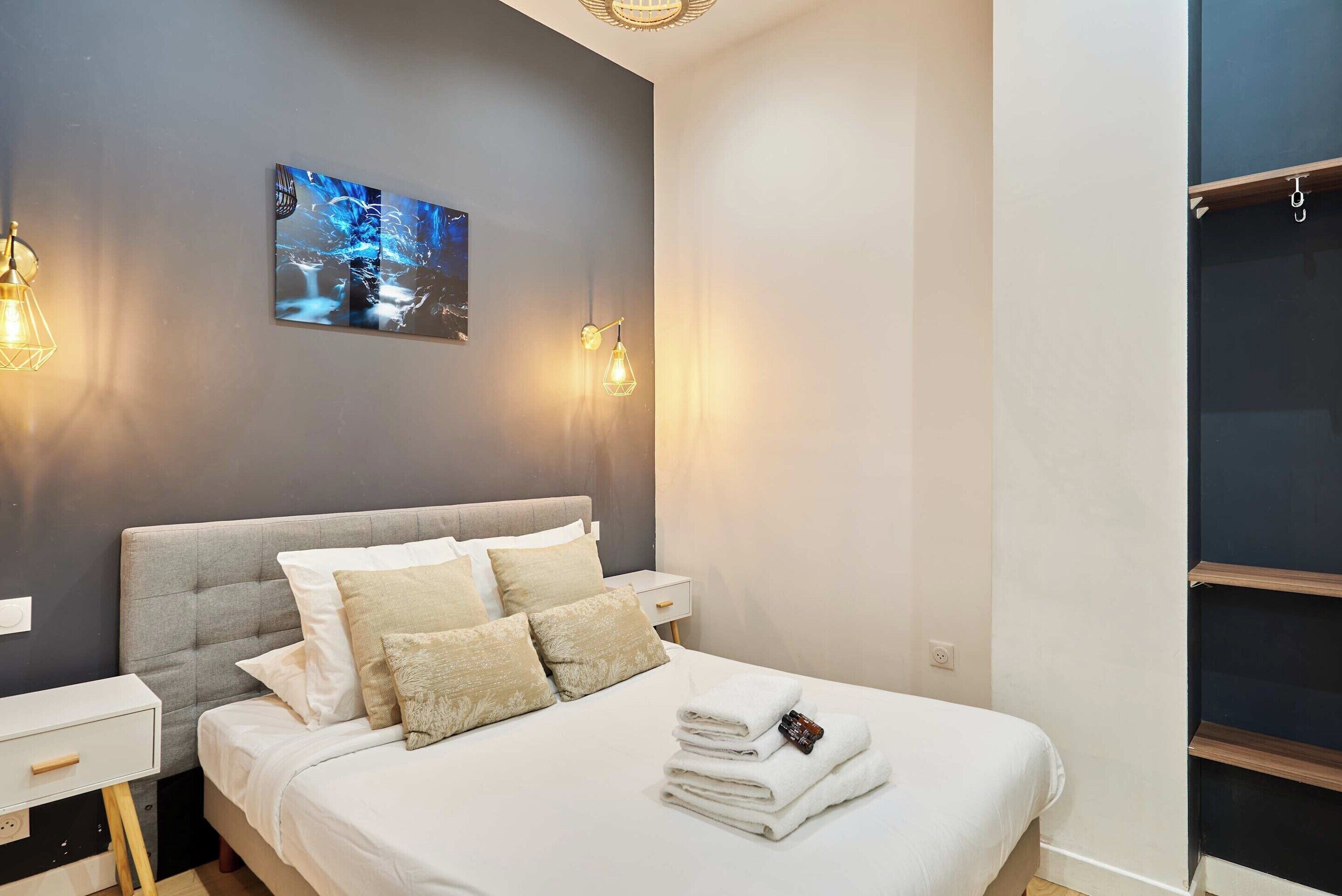 A neatly made bed with white linens and a folded towel, a grey upholstered headboard, a bedside table with a lamp, and a wall-mounted artwork.