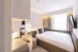 A hotel room with a large bed, a desk, a chair, a TV mounted on the wall, and a window with curtains.