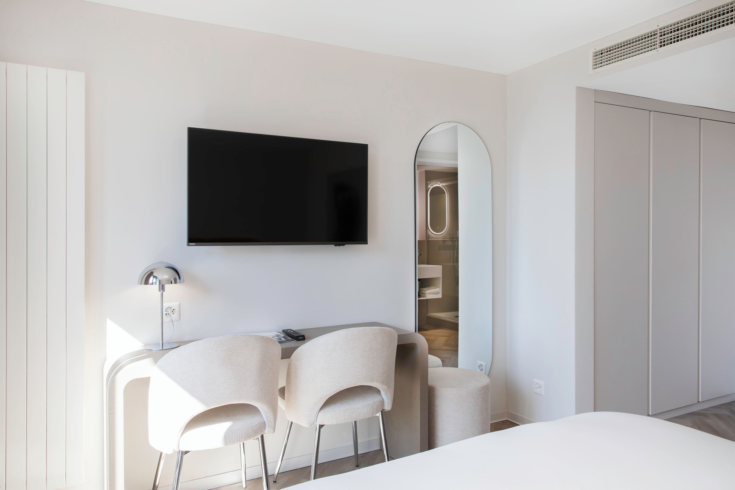 A modern hotel room with a flat-screen TV, a large mirror, two chairs, a desk, and a wardrobe.