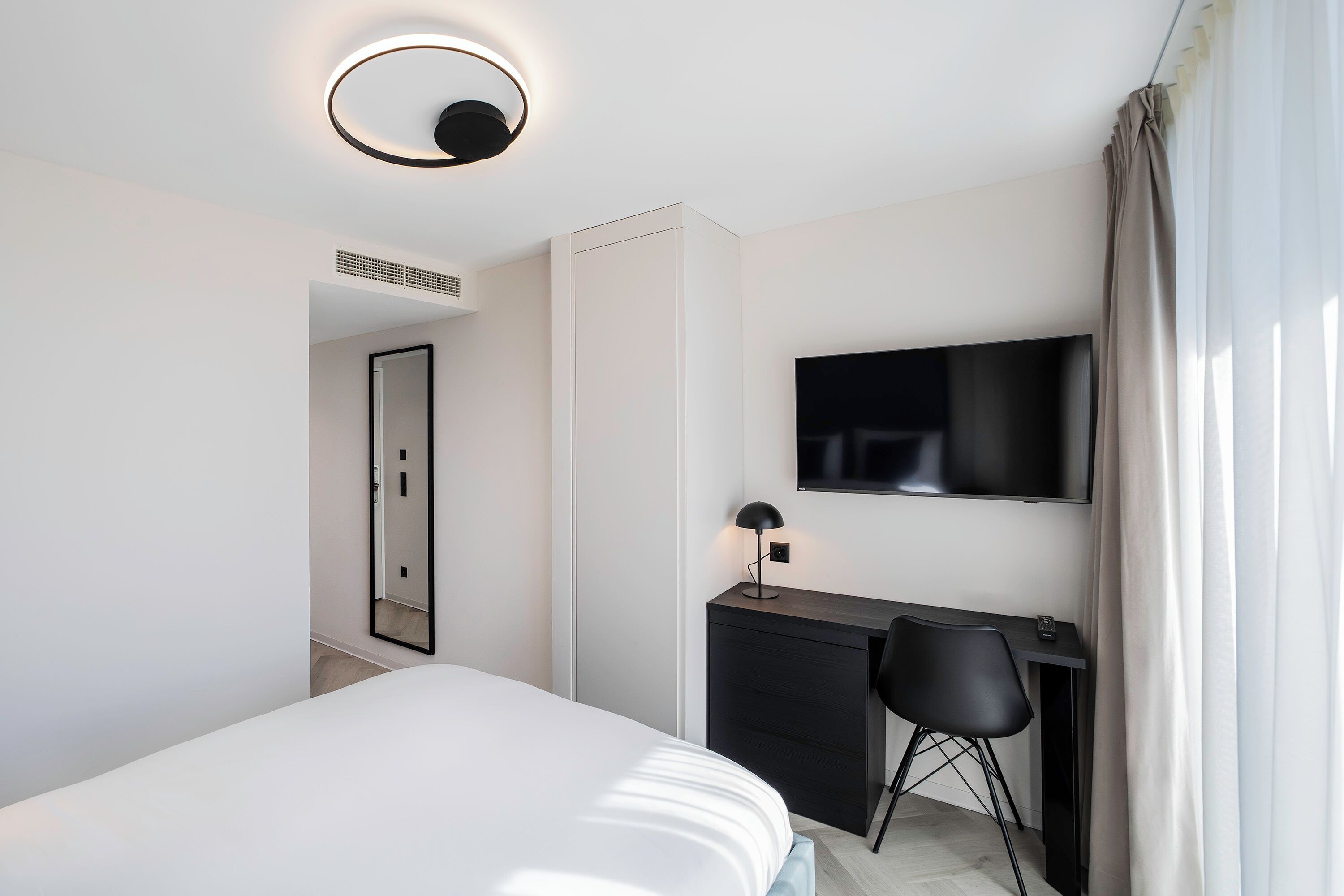 A modern hotel room with a bed, a desk, a chair, a flat-screen TV, and a ceiling light.