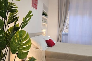 Ein ordentlich bezogenes Bett mit weißen und roten Kissen, eine Zimmerpflanze am Nachttisch und ein Fenster mit Vorhängen.