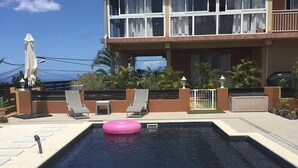 A swimming pool with a pink float, lounge chairs, and a building with large windows and a balcony. - Spacious 2-bedroom apartment in Avirons - Terrace & Parking (Les Avirons)