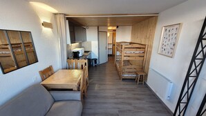 Une pièce compacte avec un lit superposé en bois, une table à manger avec des chaises, un canapé et, au fond, un coin cuisine.