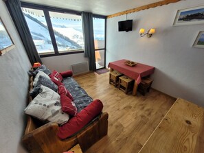 Um quarto acolhedor com um sofá de madeira, uma pequena mesa de jantar e uma televisão fixada na parede.