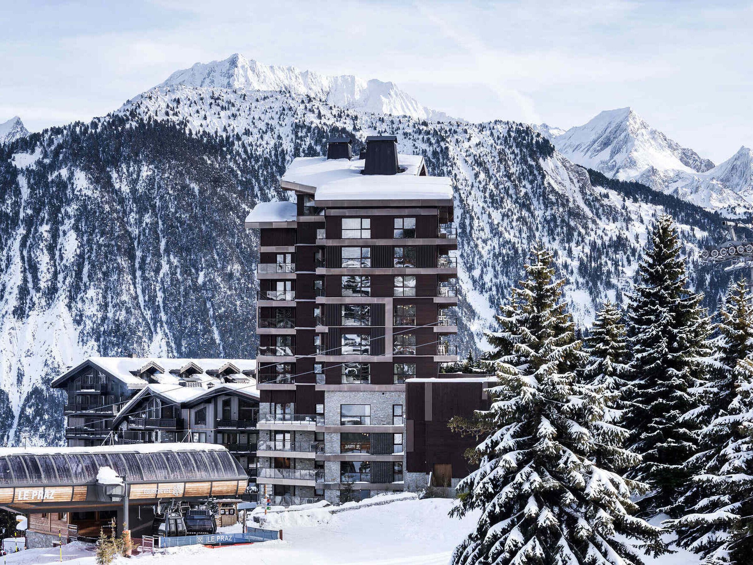 A modern multi-story hotel with a ski lift in the foreground, surrounded by snow-covered mountains.