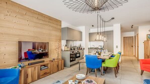 Un salon moderne avec un mur en bois, un coin repas avec des chaises colorées, une cuisine équipée d’appareils électroménagers en acier inoxydable et un luminaire au plafond.