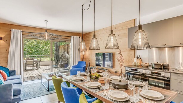 Une cuisine moderne équipée d’appareils électroménagers en acier inoxydable, un coin repas avec des chaises aux couleurs vives et une vue sur une terrasse en bois.