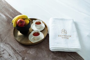 A tray with bananas, an apple, and two cups of tea, accompanied by a white towel with "KEYROCK living" embroidered on it.