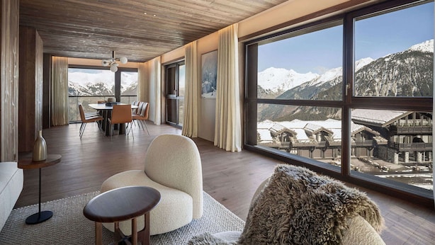 A spacious room with a wooden ceiling, large windows offering a mountain view, a dining area, and a cozy seating area with a fur throw.