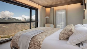 A bedroom with a large window, a bed with white linens, a wooden ceiling, and a view of snow-capped mountains.