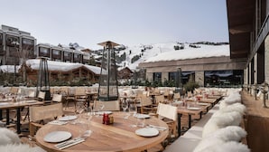 An outdoor dining area with round wooden tables and chairs, set against a backdrop of snow-covered mountains and a clear sky.