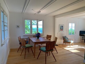 A dining area with a wooden table and chairs, large windows with a view, and a television in the corner.