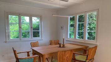 Ein Esszimmer mit Holztisch und -stühlen, großen Fenstern mit Grünpflanzen im Hintergrund und einer Deckenleuchte.