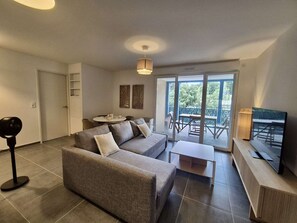 A modern living room with a grey sofa, a small coffee table, and a TV on a wooden stand. There is a balcony with a table and chairs, and a ceiling light. - Modern apartment with pool, air conditioning, and parking in Cavalaire-sur-Mer (Cavalaire-sur-Mer)