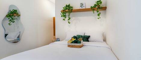 Ein ordentlich bezogenes Bett mit weißen Leinen, ein Korbwäsche auf dem Bett und eine Holzablage mit grünen Pflanzen darüber.