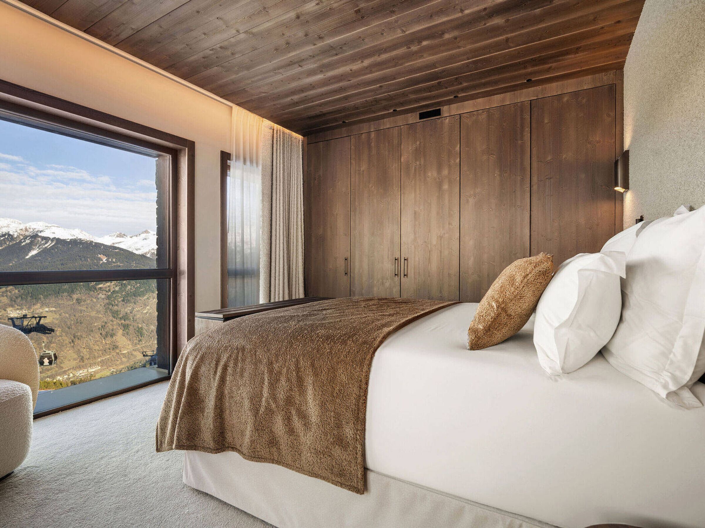 A bedroom with a large window, wooden ceiling, and a bed with white and brown bedding.
