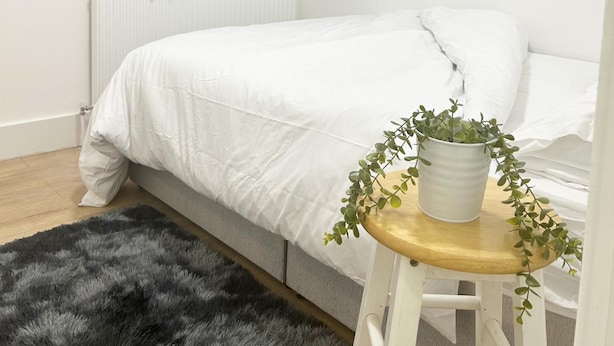 A bedroom with a bed, a radiator, a window with curtains, a grey furry rug, and a wooden stool with a potted plant.