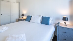 Habitación de hotel con una cama grande, dos mesitas de noche, un armario y un pequeño escritorio con lámpara.